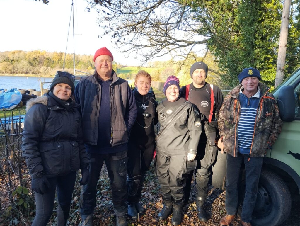 Victoria, Keith, Kerry, Clare, Adam, and Mick from the CADServation team standing side by side in front of Cransley Reservoir during the clean-up training day.